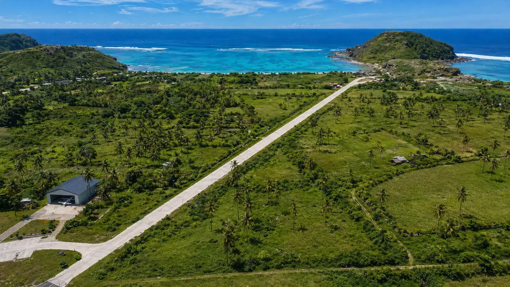 Drone aerial of the Tanjung Aan runway — hangar at the south threshold, 450 m of concrete running north toward the bay and the reef break